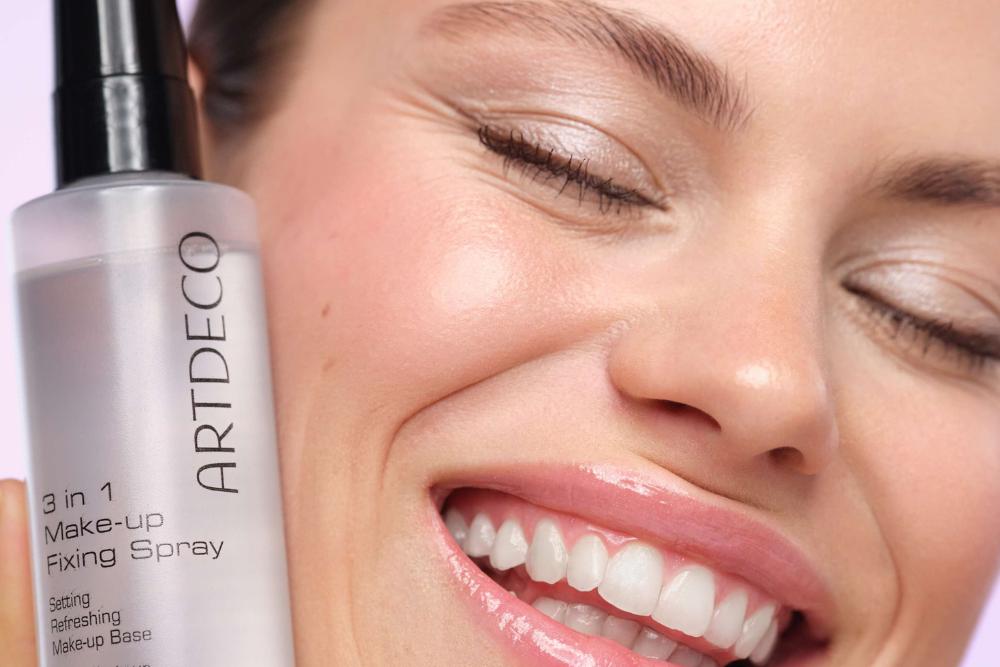 Model hält das 3 in 1 Make-up Fixing Spray an ihre Wange und lächelt dabei glücklich