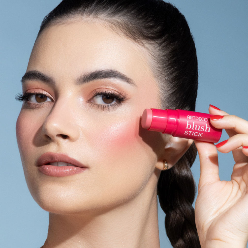 Eine Frau mit dunklen, zurückgebundenen Haaren hält sich den Blush Stick von Artdeco an die linke Wange
