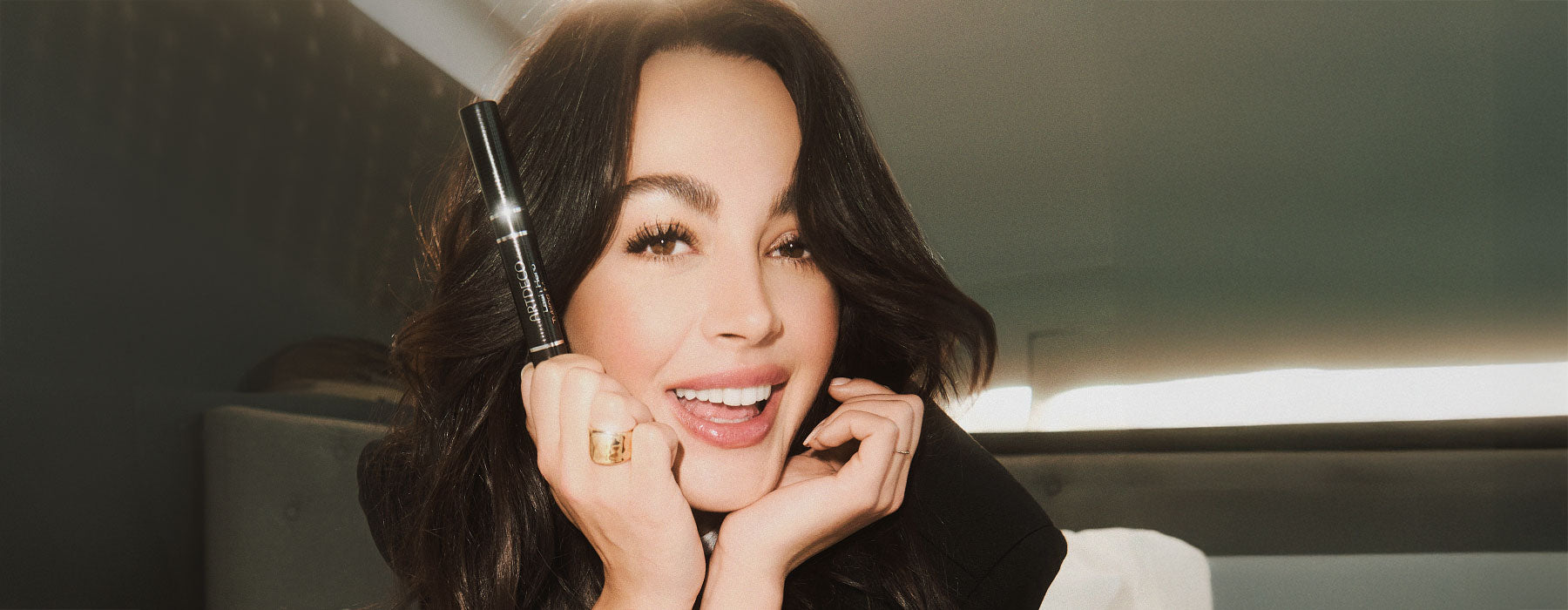 Brünette Model lächelt und die Lash Hero Tubing Mascara dabei in der Hand hält