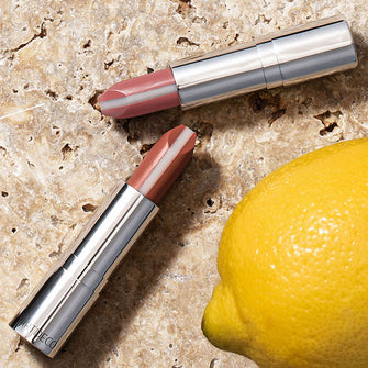 Zwei Hydra Care Lipsticks liegen auf einem Steinhintergrund, umgeben von einer sommerlichen Zitrone