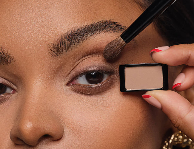 Model hält den neutralen Lidschatten in der Hand und zeigt die Anwendung am Auge mit einem fluffigen Lidschatten-Pinsel