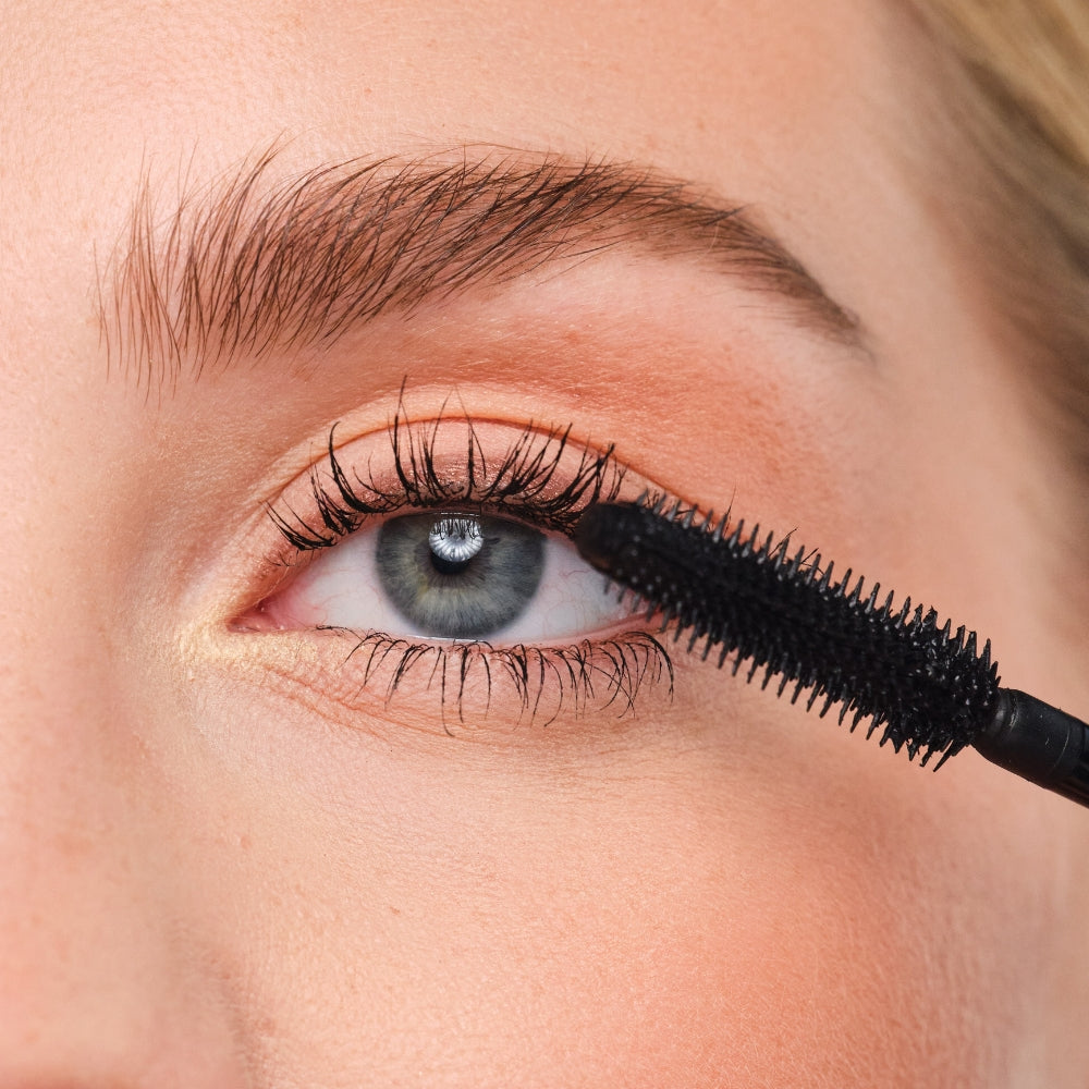 Close-up eines Auges mit einer Mascara-Bürste, die vor dem Auge gehalten wird. Die Wimpern sind deutlich sichtbar.