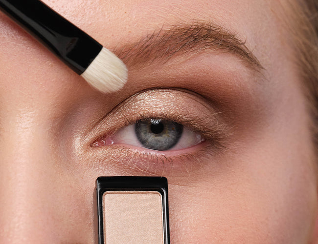 Close-up auf das Auge des Models, auf welchem Lidschatten in einem Champagner-Ton in Augeninnenwinkel aufgetragen wird