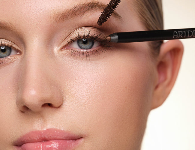 Close-up auf das fertig geschminkte Auge des Models, an welches eine braune Mascara und ein brauner Eyeliner gleichzeitig gehalten werden