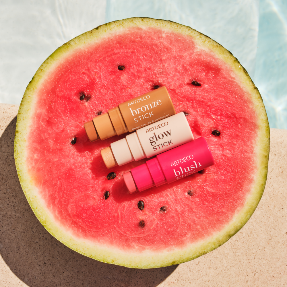 Bronze Stick, Blush Stick und Glow Stick liegen gemeinsam auf einer halbieren Melone an einem Pool.