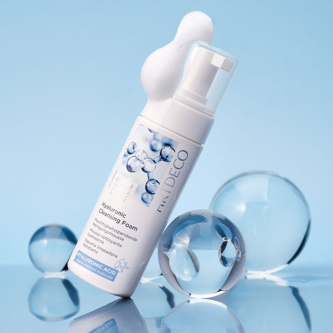 Der Hyaluronic Cleansing Foam vor einem hellblauen Hintergrund umgeben von verschieden großen Glaskugeln. Auf dem Deckel ist etwas Schaum zu sehen.