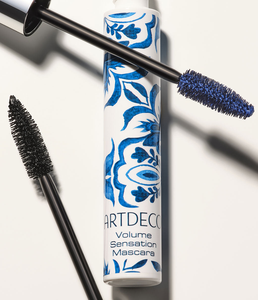 Artdeco Volume Sensation in Weiß mit blauem Azulejo-Design. Zwei Bürstenstäbe mit unterschiedlicher Textur, einer mit schwarzer und einer mit blauer Mascara, liegen auf einem hellen Hintergrund