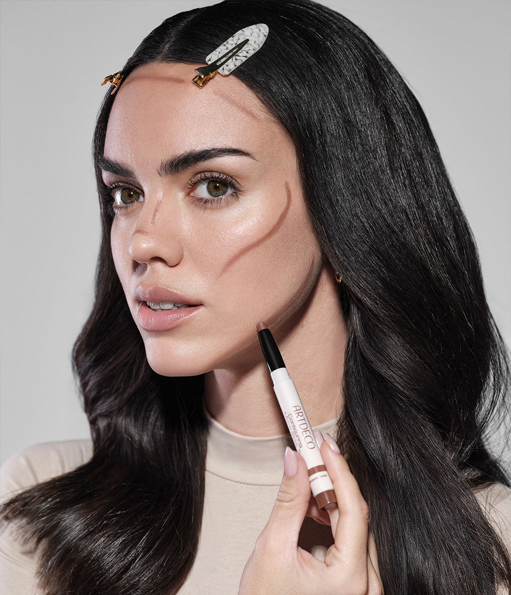 Brünettes Model hält den Contouring Micro Stick in ihrer Hand. Dieser wurde bereits auf Stirn, Nase und Wangen aufgetragen, aber noch nichts verblendet.