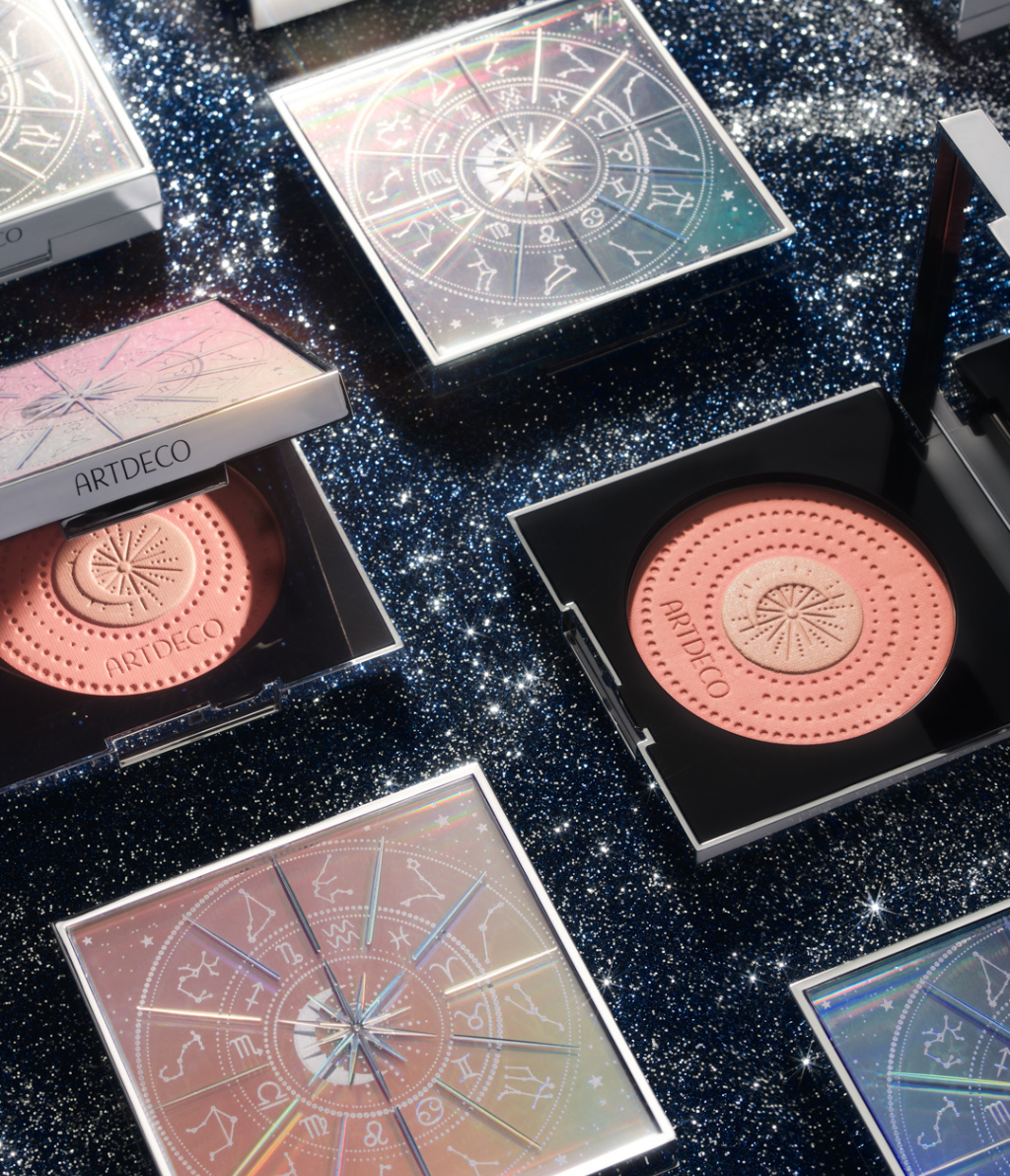Mehrere Galactic Blush Couture liegen entweder geöffnet der geschlossen auf einem blauen Glitzer-Hintergrund. Das limitierte Design und die hochwertige Prägung sind gut erkennbar.