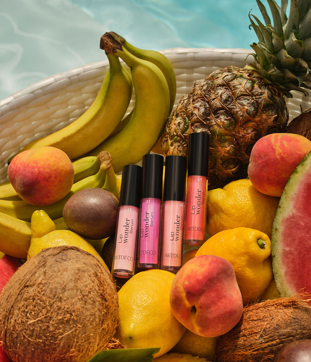 Lip Wonder Serum in vier sommerlichen Farben, platziert in einem Korb voller buntem Obst.