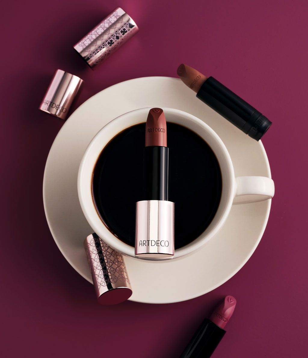 Der Couture Lipstick im zarten Rosé-Design liegt senkrecht auf einer Tasse, die umgeben von weiteren Lippenstiften ist