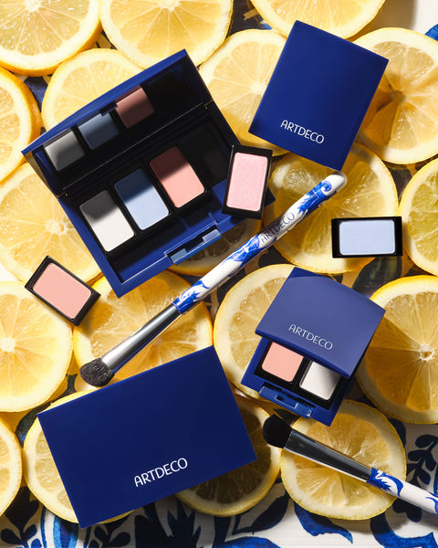 Verschiedene Beauty Boxen im limitierten blauen Design werden auf einem Hintergrund mit frischen Zitronenschreiben präsentiert. Ergänzt wird die Szene durch einzelne Lidschatten in Blau- und Rosatönen sowie einen Pinsel mit Azulejo-Fliesen-Design.