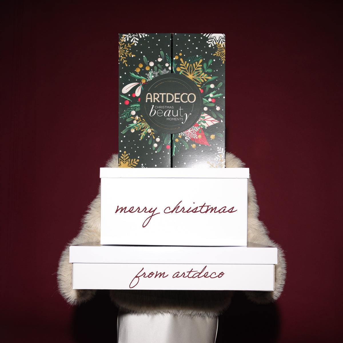 Eine Person hält drei übereinandergestapelte Geschenkboxen vor sich. Oben steht der grüne, weihnachtlich verzierte ARTDECO Adventskalender, darunter zwei weiße Boxen mit der roten Aufschrift „merry christmas“ und „from artdeco“.