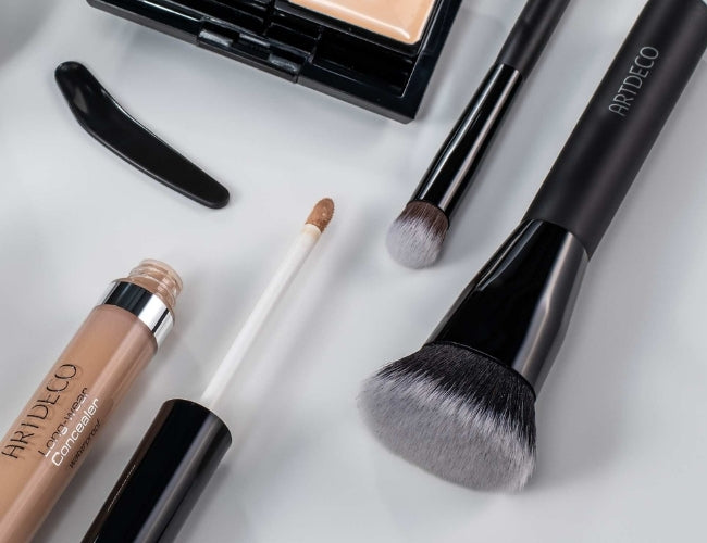 Verschiedene langanhaltende ARTDECO Make-Up Produkte und Pinsel platziert auf grauem Hintergrund.