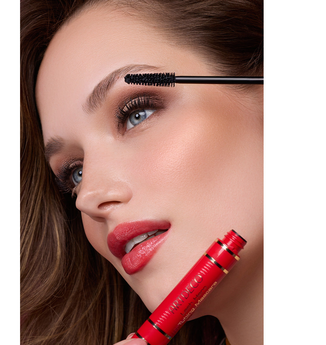 Model steht seitlich zur Kamera, während sie die Lash Hero Tubing Mascara im limitierten roten Design aus der Whispers of the Silk Road-Kollektion aufträgt.