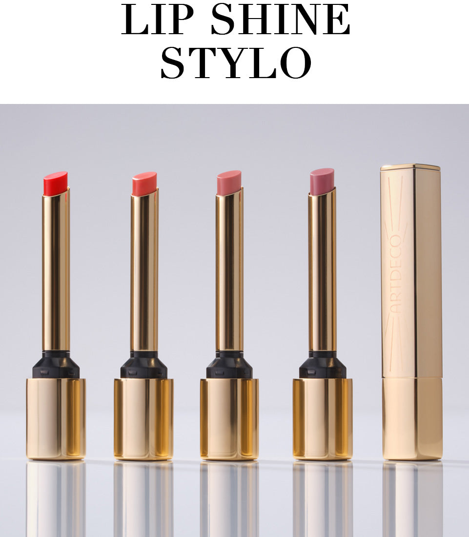 Alle vier Lip Shine Stylo stehen geöffnet aufgereiht auf einem neutralen Hintergrund. Ein fünfter Lip Shine Stylo steht geschlossen daneben. Die filigrane, goldene Verpackung vermittelt Hochwertigkeit des Produktes. Zudem steht in Großbuchstaben der Name des Produktes »Lip Shine Stylo«.