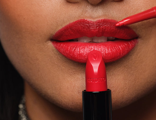 Close-up auf die geschminkten Lippen des Models an welche ein roter Lipliner und roter Lippenstift gehalten werde