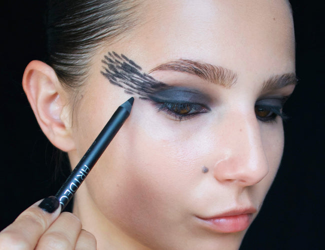 Das Model zeichnet mit einem schwarzen Eyeliner unterschiedlich lange Striche, welche am Schluss eine Art Flügelform ergeben. 