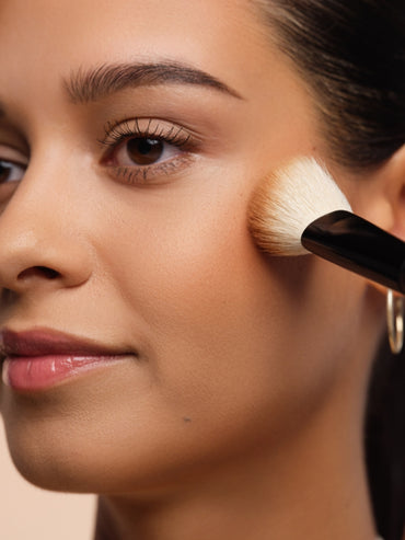 Model trägt mit einem weichen Pinsel Bronzer auf die Wangenknochen auf um dem Gesicht eine natürliche Wärme und Kontur zu verleihen.