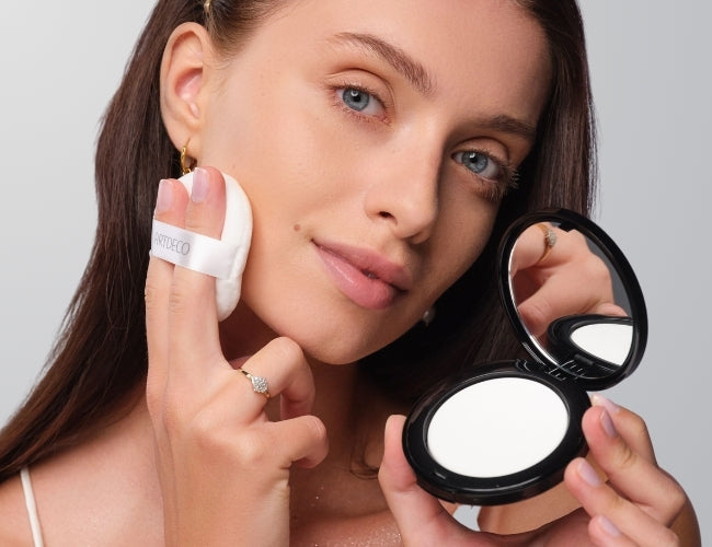 Model mit dunklen Haaren trägt mit einer Quaste transparentes Puder gleichmäßig auf ihrem Gesicht auf.