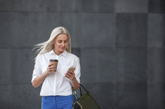 Frau mit langen blonden Haaren hält Kaffeebecher in der Hand und schaut auf ihr Handy