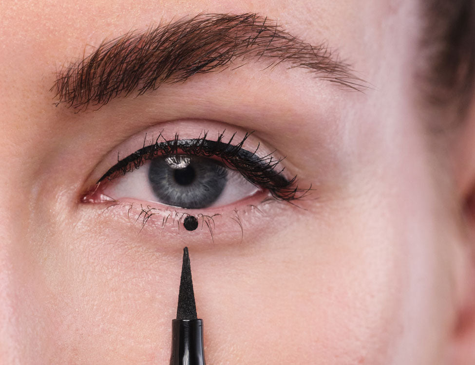 An ein Auge mit gezeichnetem Eyeliner und kreativem Punkt am unteren Wimpernkranz wird eine Eyeliner -Spitze gehalten. 