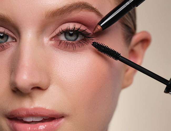 Close-up auf das fertig geschminkte Auge des Models an welches eine schwarze Mascara und ein kupferfarbener Lidschattenstift gleichzeitig gehalten werden