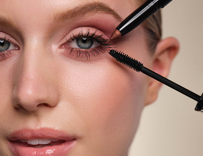 Close-up auf das fertig geschminkte Auge des Models an welches eine schwarze Mascara und ein kupferfarbener Lidschattenstift gleichzeitig gehalten werden
