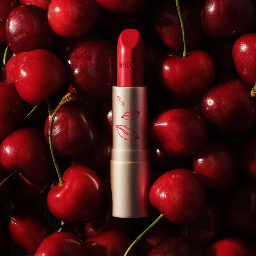 Ein Perfect Color Lipstick in roter Farbe liegt ohne Deckel auf Kirschen